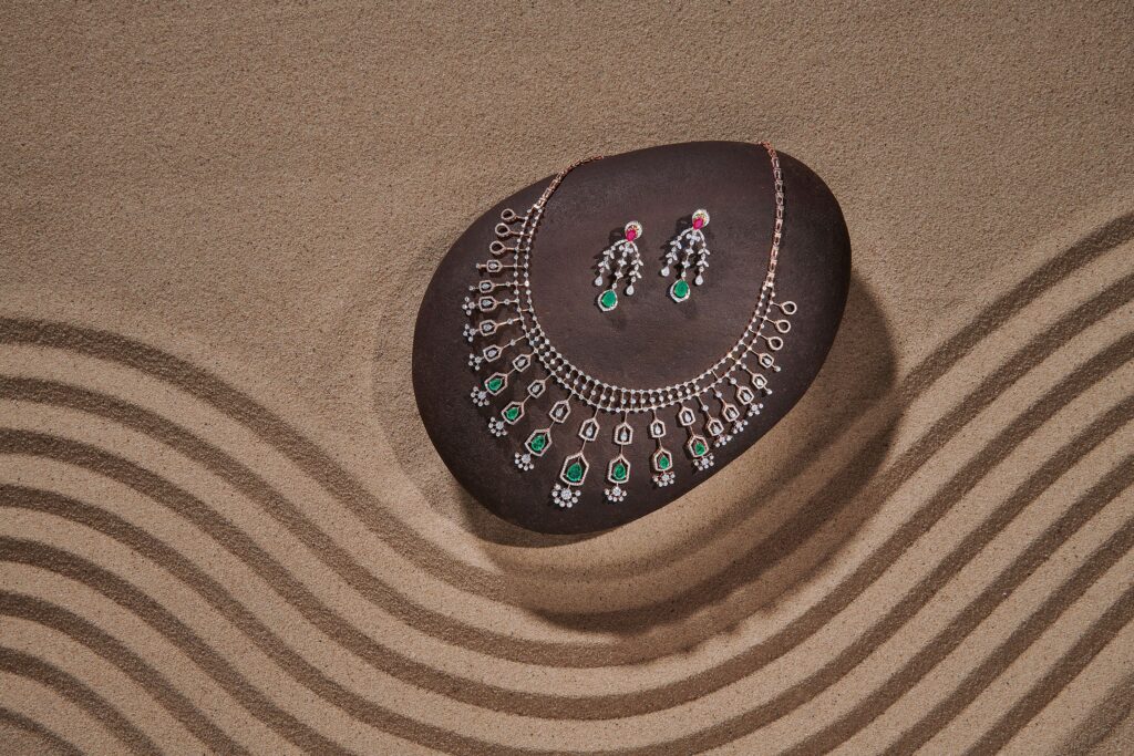 Bhima diamond necklace and earrings set displayed on a brown stone surrounded by sand-art patterns.
