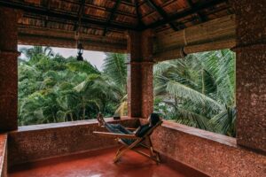 A person relaxing on a wooden lounge chair in a rustic open terrace surrounded by lush green palm trees at Poothali Homestay.