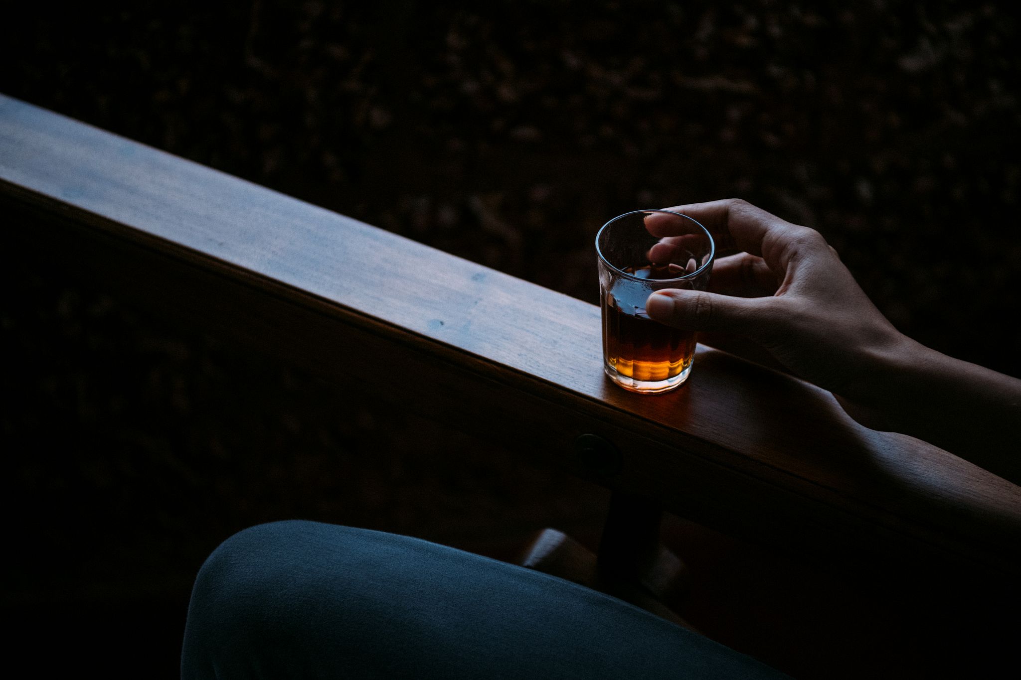 A hand holding a glass of amber-colored drink while resting on a wooden armrest.