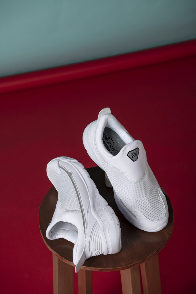 A pair of stylish white slip-on sneakers from the brand “Puca,†displayed on a wooden stool with a bold red and teal background.