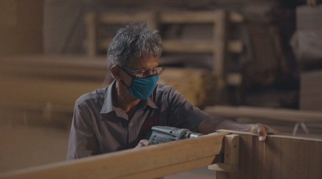 Carpenter wearing a mask using a power drill on wooden furniture.