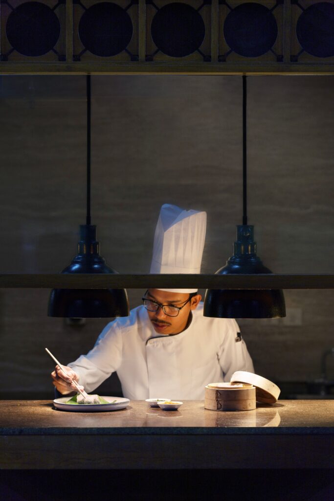 A Taj Chia Kutir chef in uniform plating food under warm pendant lights in the kitchen.