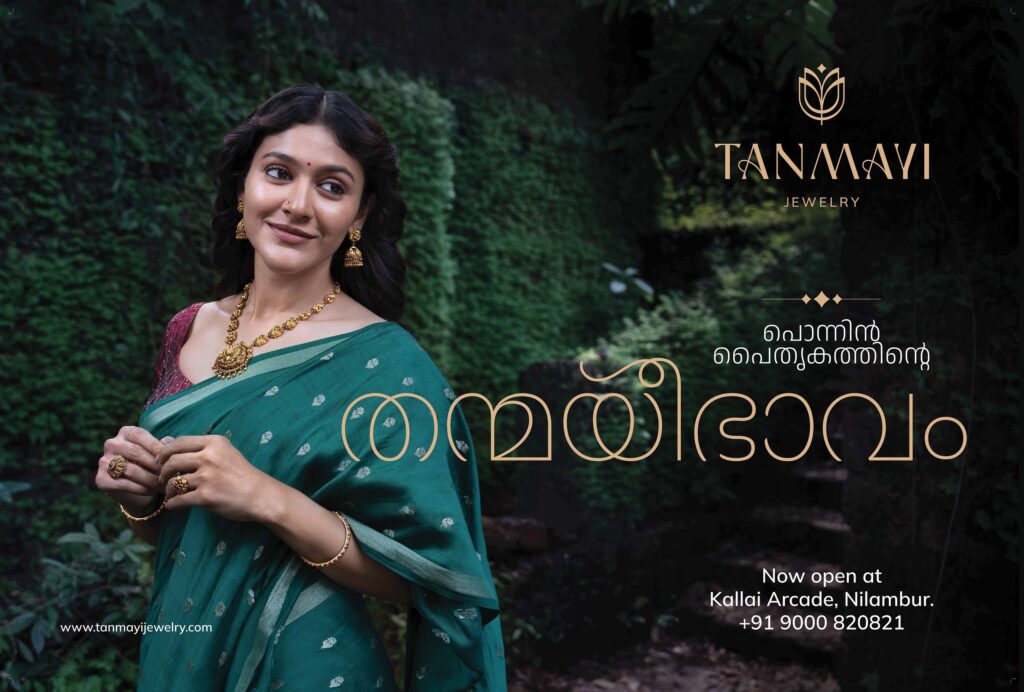 A woman in a green saree adorned with gold jewelry, smiling gracefully against a lush green backdrop, showcasing Thanmayi Jewellery.