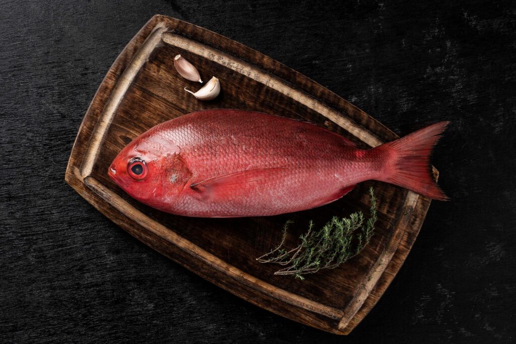 Whole raw red fish on a wooden board with garlic and herbs.