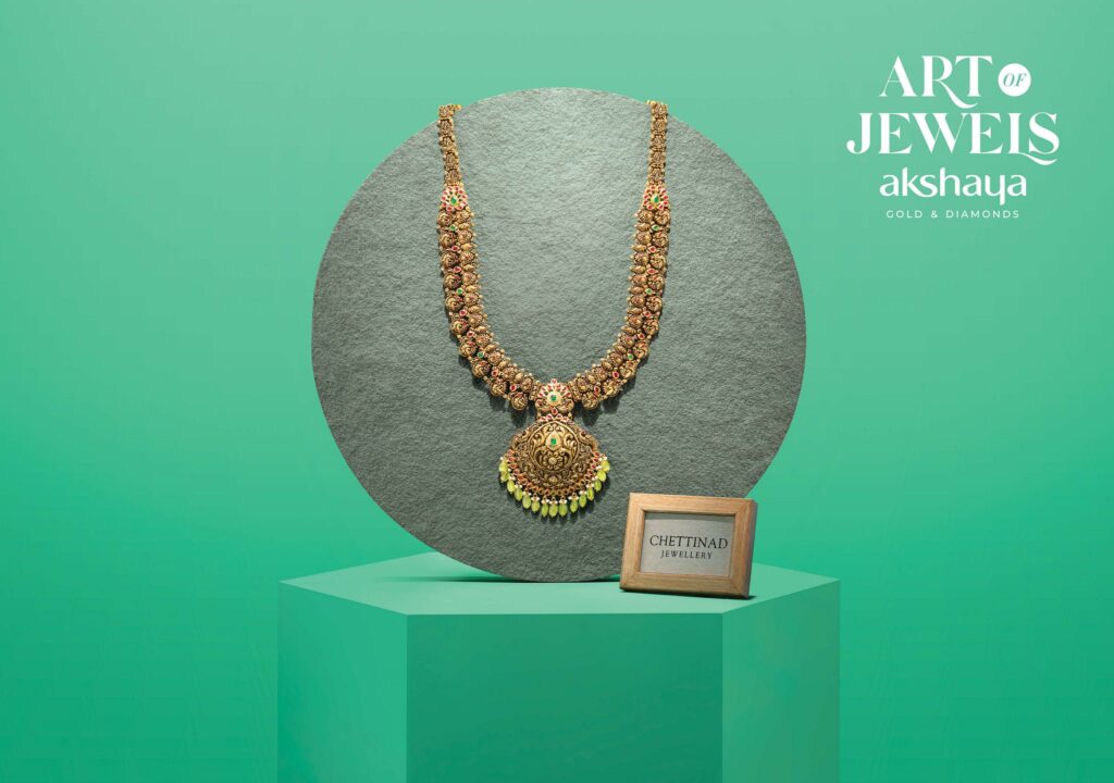 Akshaya Gold & Diamonds Art of Jewels campaign: a traditional gold Chettinad necklace displayed on a pedestal against a green background.