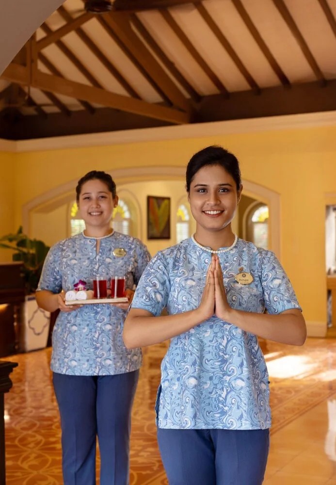 Two hospitality staff members in matching blue patterned uniforms stand inside a warm, well-lit interior